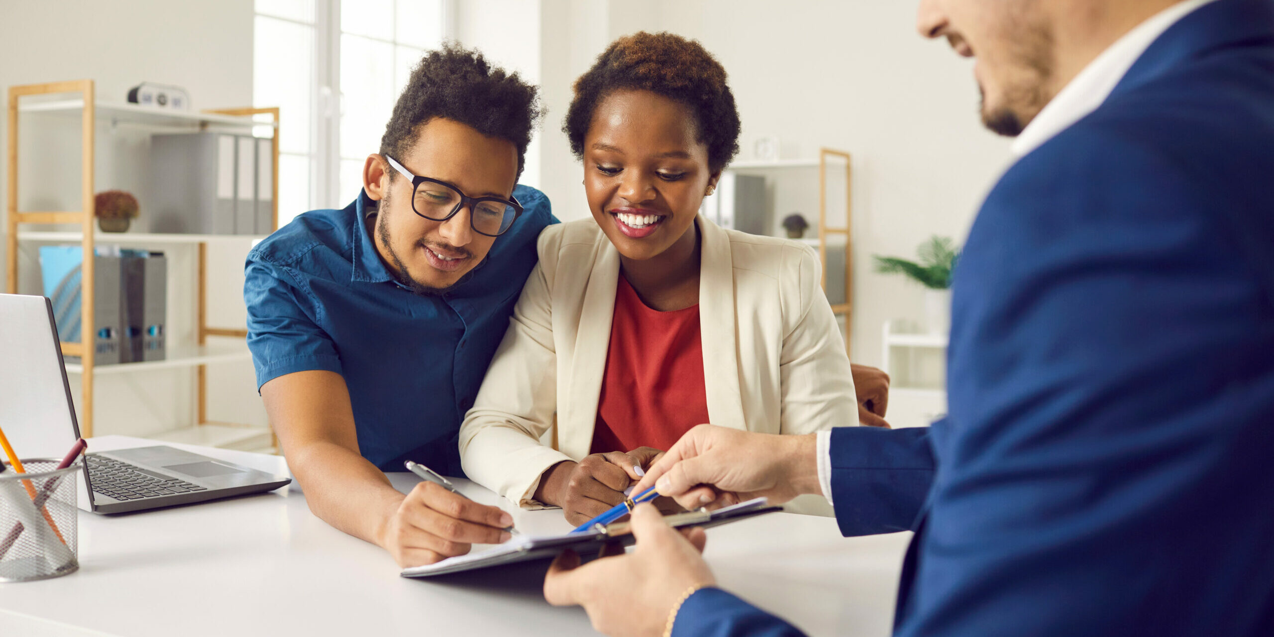 Happy family signing contract after consulting with real estate agent or mortgage broker. Mixed race couple at office desk together putting signatures on contract of sale or notary rent agreement